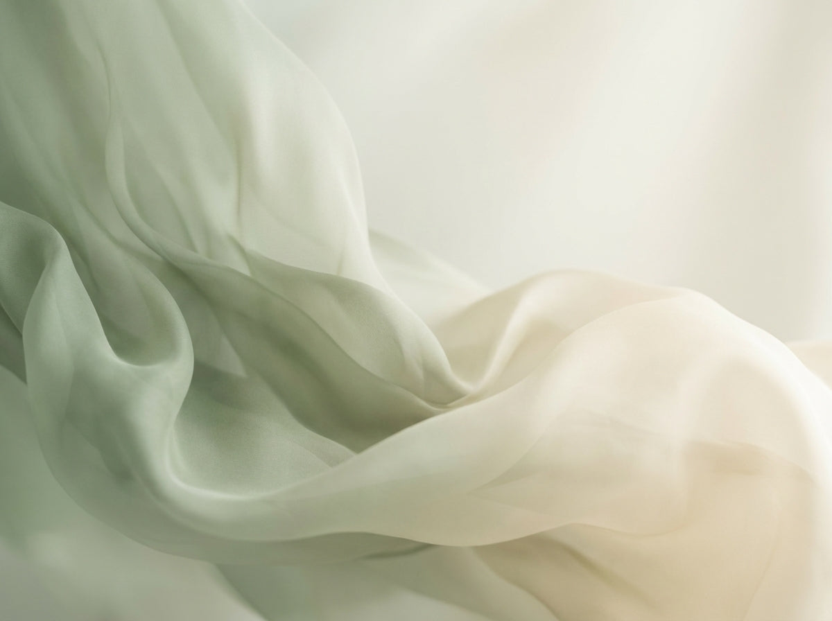 Soft draped fabric in a pale sage green and cream tone, shown with flowing folds and a lightweight, semi-sheer texture against a softly lit neutral background.